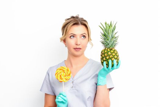 Young caucasian female doctor nutritionist holding fresh pineapple with lollipop over white background