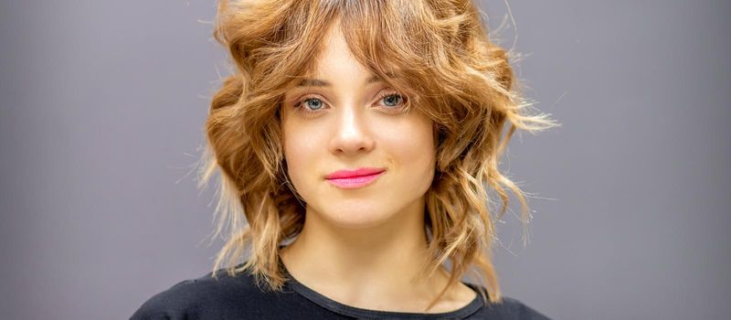 Portrait of a beautiful young caucasian red-haired woman with short wavy hairstyle smiling and looking at camera on dark gray background with copy space