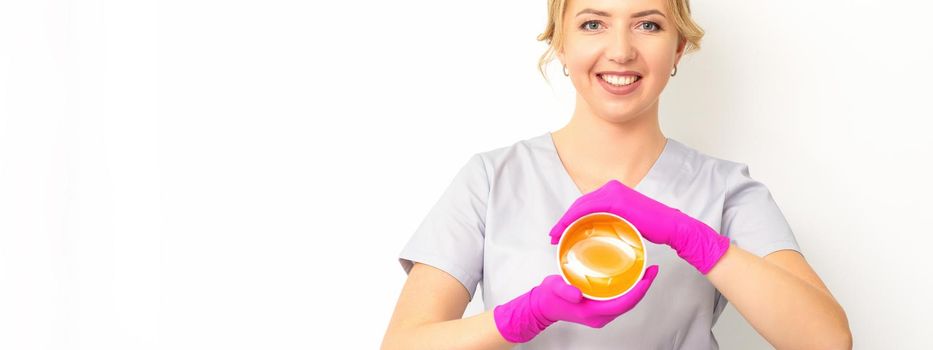 Portrait of a female caucasian beautician holding a jar of sugar paste for sugaring wearing pink gloves on white background