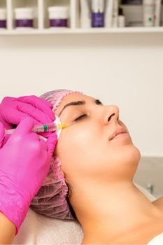 The young white woman is getting rejuvenating facial injections with hyaluronic acid on the eye in a beauty clinic