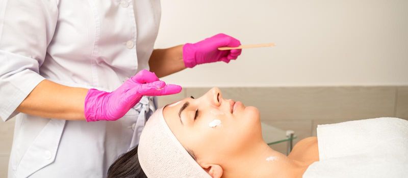 Cosmetologist with gloved hands applies a moisturizing mask with peeling cream on the female face. Facial cosmetology treatment. Procedures for facial care