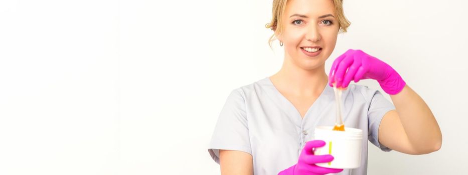 Portrait of a female caucasian beautician holding a jar of sugar paste for sugaring wearing pink gloves on white background