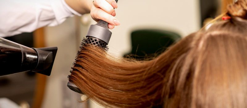 A hairdresser is drying long brown hair with a hairdryer and round brush in a beauty salon