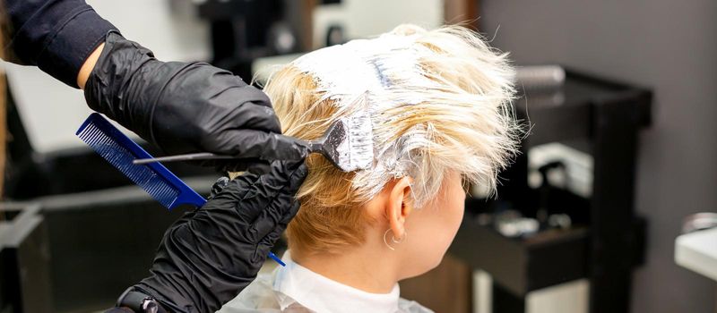 Coloring white hair with hair dye and brush by hands of hairstylist for the young caucasian blonde woman at a hair salon, close up