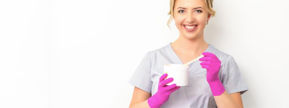 Portrait of a female caucasian beautician holding a jar of sugar paste for sugaring wearing pink gloves on white background