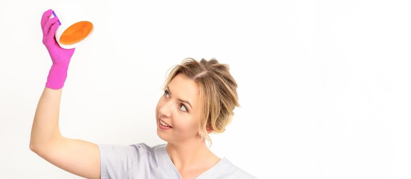 Portrait of a female caucasian beautician holding a jar of sugar paste for sugaring wearing pink gloves on white background