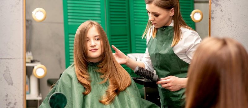 Professional hair care. Young female red-haired with long hair receiving hairstyling in a beauty salon.