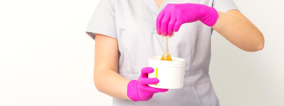 Portrait of a female caucasian beautician holding a jar of sugar paste for sugaring wearing pink gloves on white background