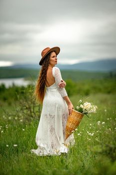 A middle-aged woman in a white dress and brown hat stands on a green field and holds a basket in her hands with a large bouquet of daisies. In the background there are mountains and a lake