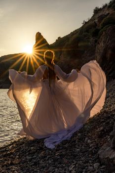 A mysterious female silhouette with long braids stands on the sea beach with mountain views, Sunset rays shine on a woman. Throws up a long white dress, a divine sunset