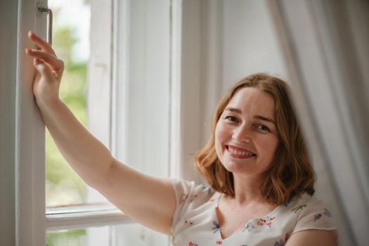 A middle-aged woman in a cream dress sits mysteriously and looks out the window on the windowsill. Green trees outside