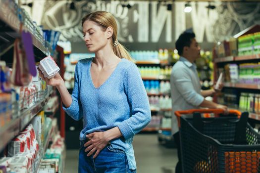 A young woman stands in a supermarket in the household department and chooses and buys feminine hygiene products on the shelf, pads. Holds the stomach, feels discomfort and pain during menstruation.