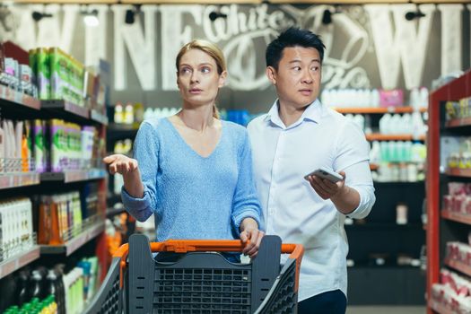 Confused young international couple, woman and Asian man walking between rows with grocery cart and phone in handsin supermarket. They spread their hands, looking for goods.
