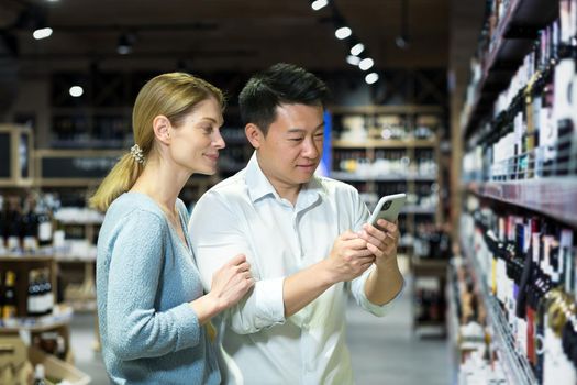 Young international beautiful couple, Asian man and blonde woman choose wine in liquor department in supermarket, use phone with app.