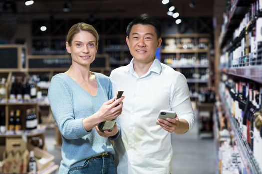 A young interracical family, an Asian man and a woman, are buying wine. They are standing in the alcohol section of the supermarket, looking at the camera. They are holding a bottle and a phone.