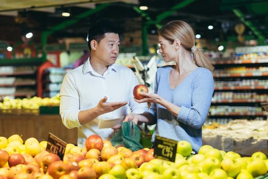 A young international couple, a woman and an Asian man, are choosing fruits in a supermarket, deciding which apples to buy. They are standing in the fruit and vegetable department with a plastic bag.