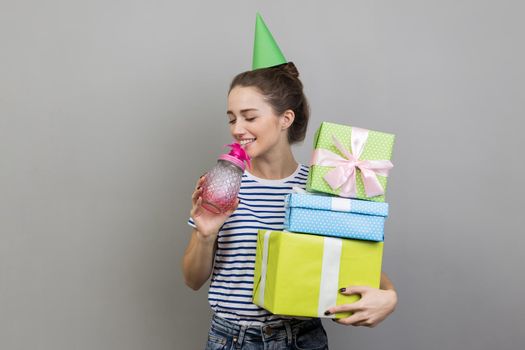 Portrait of festive positive woman wearing striped T-shirt and party cone, holding stack of present boxes, drinking cocktail, enjoying birthday party. Indoor studio shot isolated on gray background.