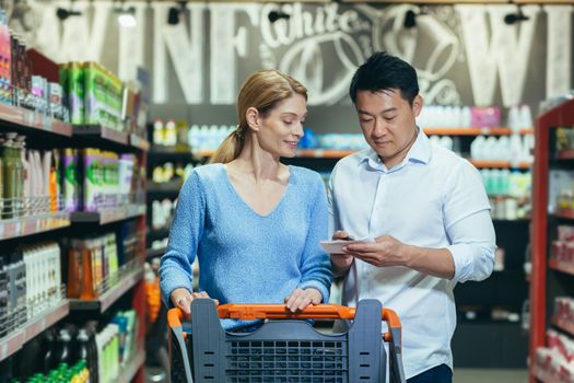 A young international couple, a woman and an Asian man, are standing in a supermarket in the department of household chemicals with a cart, holding a list of products in their hands, reading.