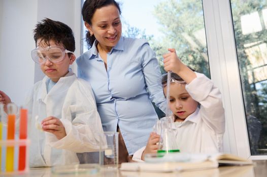 Happy school kids in protective eyewears and white lab coat, with a scientist doing science experiments in the chemical laboratory. Learning chemistry. Back to school. Open to new knowledges