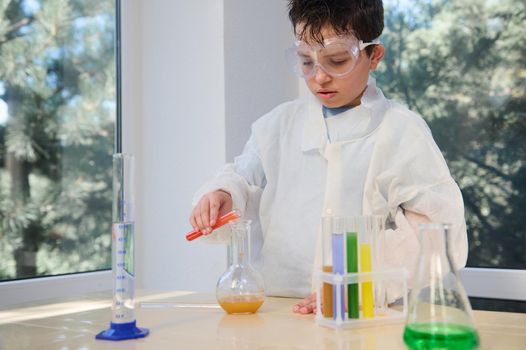 Elementary student boy in a white lab coat and safety goggles, conducting a chemical experiment, pouring liquid from test tube into a lab flask, learning Chemistry in science class .Education concept