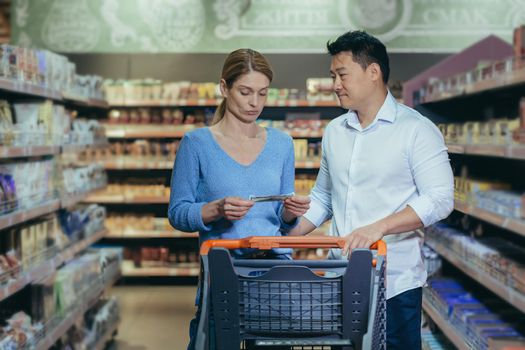 A young interracical couple, a woman and an Asian man, are standing in a supermarket in the department of household chemicals with a cart, holding a list of products in their hands, reading.