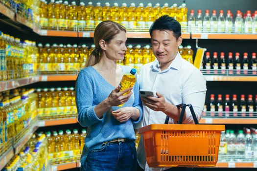 Young international family, Woman and asian man choose oil in supermarket. They hold a bottle of oil, a basket and a phone. They are looking for the brand, composition of the oil in the search engine.