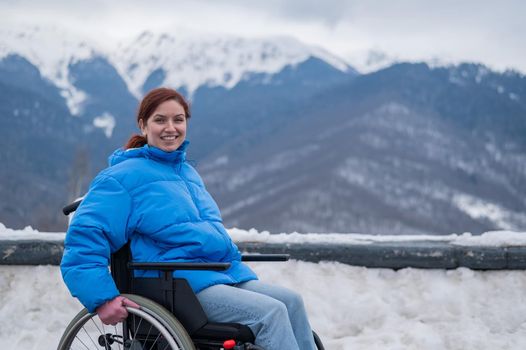 Smiling woman dressed in a blue coat sits in a wheelchair on a point view looks at high mountains