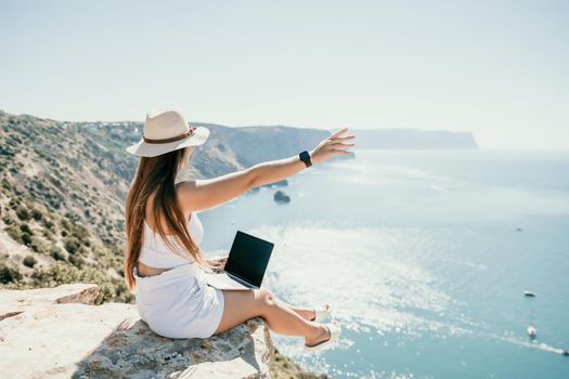 Successful business woman in yellow hat working on laptop by the sea. Pretty lady typing on computer at summer day outdoors. Freelance, travel and holidays concept.