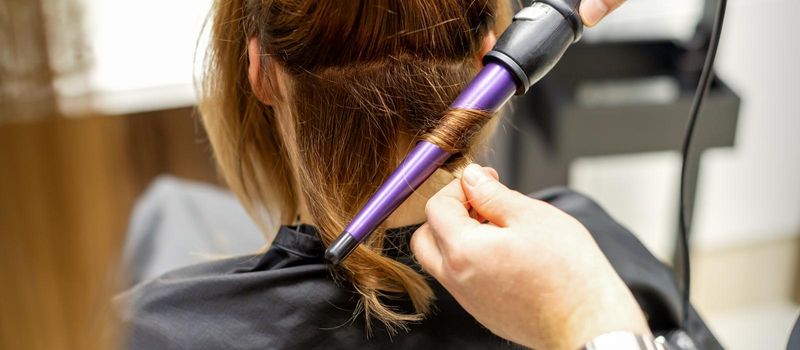 The hairstylist makes curls hairstyle of long brown hair with the curling iron in hairdresser salon, close up