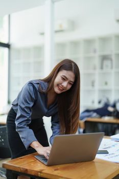 Portrait of an Asian female employee standing at a computer with a smiling face as she works the next morning.