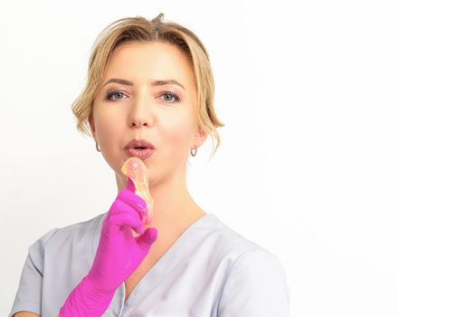 Young caucasian female beautician holding wax paste on forefinger blowing on him wearing pink gloves against a white background. Epilation concept