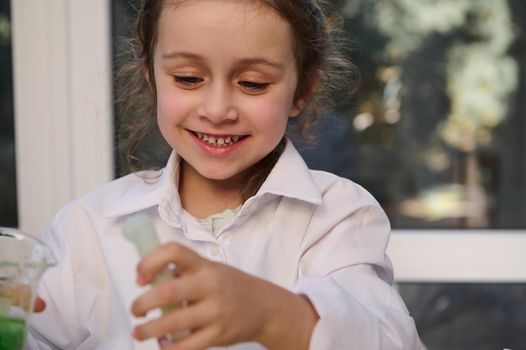 Portrait of a Caucasian mischievous little girl in lab coat, holding a laboratory test tube and a flask with chemicals and reagents, watching the taking place chemical reaction in the Chemistry class