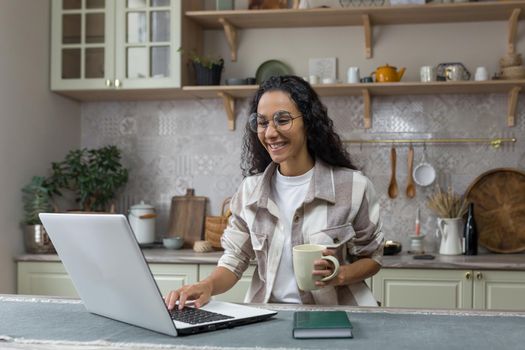 Young beautiful hispanic woman with curly hair working at home using laptop, woman in kitchen smiling and happy remote work freelancer.