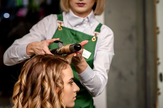 The female hairdresser is curling hair for a brown-haired young caucasian woman in a beauty salon