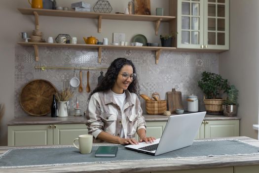 Young beautiful hispanic woman with curly hair working at home using laptop, woman in kitchen smiling and happy remote work freelancer.
