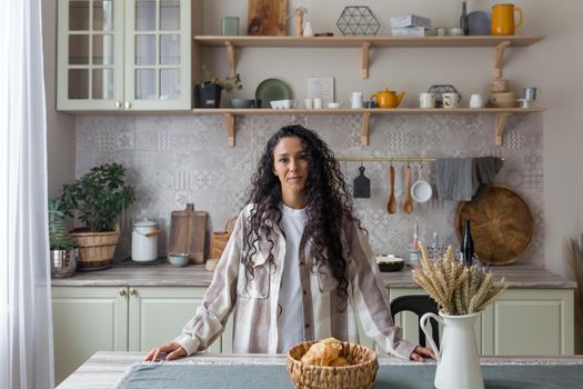 Portrait of happy hispanic woman at home in kitchen, woman with curly hair smiling and looking at camera.