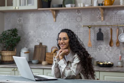 Portrait of young beautiful hispanic woman at home, female freelancer working remotely using laptop, looking at camera and smiling in glasses and curly hair in kitchen
