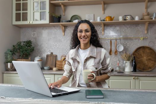 Portrait of young beautiful hispanic woman at home, female freelancer working remotely using laptop, looking at camera and smiling in glasses and curly hair in kitchen.