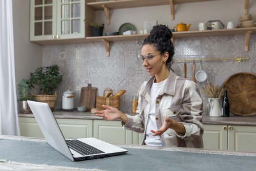Young beautiful woman in glasses and curly hair talking on video call, Hispanic woman using laptop for remote communication at home in kitchen.