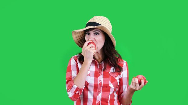 funny, smiling female farmer in checkered shirt and hat, holding in hands two large sour apples, eats, bites them, wrinkles her face, on green background in studio. Healthy food to your table. High quality photo