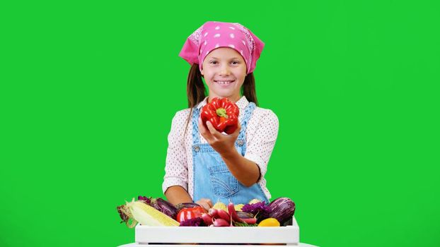 cute, smiling, little farmer girl shows a box with different fresh vegetables, harvest. on green background in studio. Healthy food to your table, Healthy nutrition. High quality photo