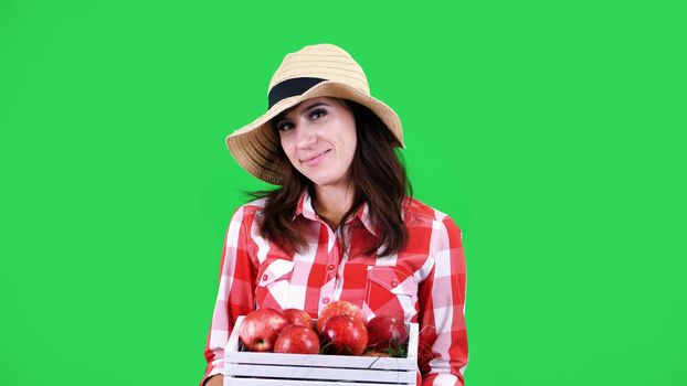 portrait of smiling female farmer in checkered shirt and hat, holding a wooden box with red ripe organic apples, on green background in studio. Healthy food to your table. High quality photo