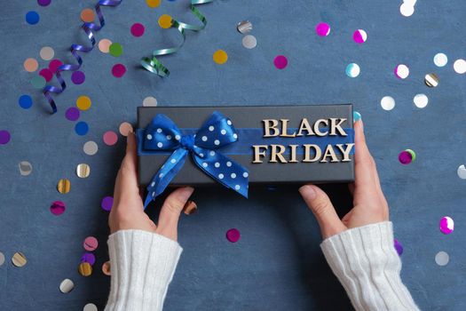 Black Friday text with gift in female hands and festive tinsel flat lay on dark cement background.