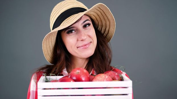 portrait of beautiful smiling female farmer in plaid shirt, gloves and hat holding white wooden box with red ripe organic apples, on gray background, in studio,. High quality photo