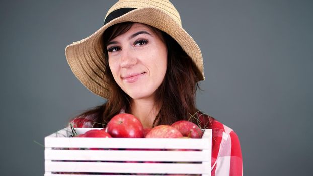 portrait of beautiful smiling female farmer in plaid shirt, gloves and hat holding white wooden box with red ripe organic apples, on gray background, in studio,. High quality photo