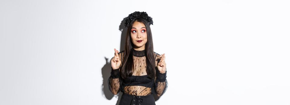 Hopeful cute asian woman in gothic lace dress and black wreath, looking upper left corner and cross fingers for good luck, making wish or praying, standing over white background.