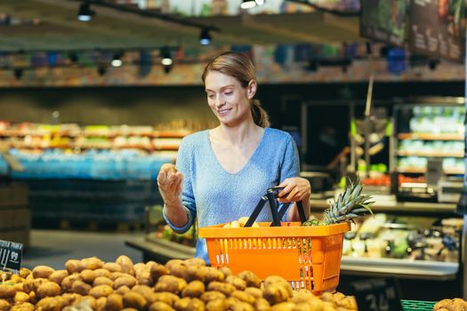 Young beautiful blonde woman in a blue sweater with a basket of fruits in her hand. Standing at the vegetable section, choosing potatoes.