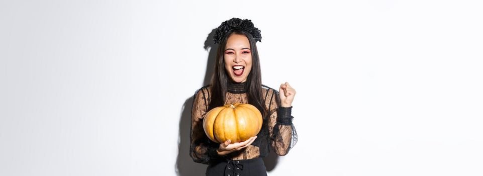 Image of excited beautiful asian woman celebrating halloween, wearing witch outfit and holding pumpkin, yelling with rejoice.