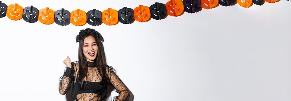 Image of happy asian woman in witch costume having fun on halloween party, saying yes cheerful, standing against pumpkin banners.