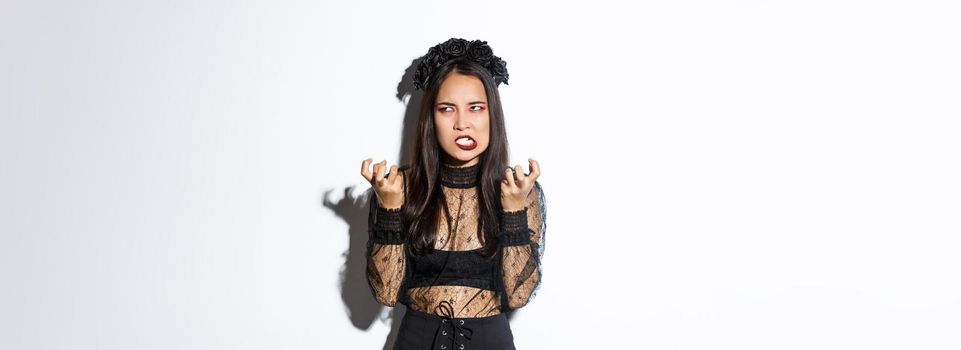 Image of annoyed and bothered asian woman looking mad, losing temper, clenching fists mad and rolling eyes, standing in evil witch costume on halloween, white background.